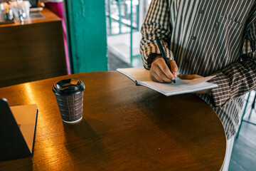 Close-up of woman writing in open notebook with black pen while standing at wooden cafe table with...