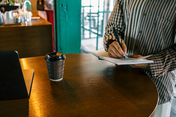 Close-up of woman writing in open notebook with black pen while standing at wooden cafe table with...