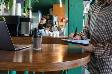 Person writing in notebook with black pen while standing at wooden cafe table with coffee cup and laptop in background