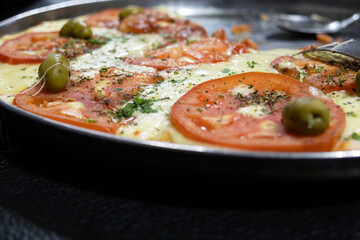A pizza with tomato slices, olives and green seasoning sprinkled over melted mozzarella