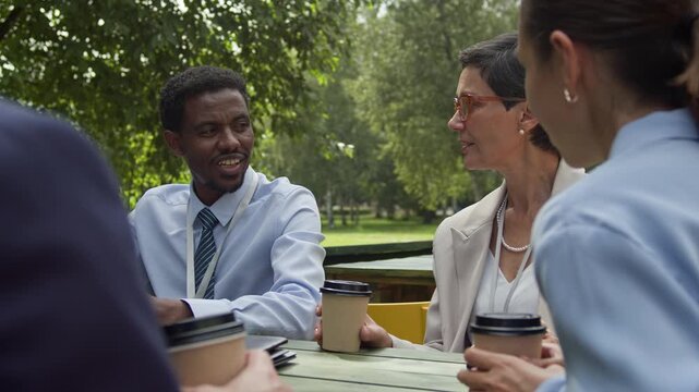 Young African American businessman in formalwear sitting by table in outdoor cafe among his female colleagues and looking at mature delegate while speaking about his native tongue