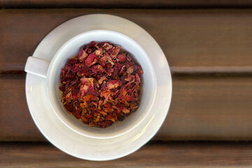 A top-down view of a white ceramic cup filled with dried rose petals, placed on a matching saucer on a wooden surface. Concept of natural beauty, herbal tea, and wellness.