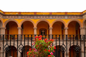 Red flowers in foreground with wrought iron railing and arched colonial architecture in former...
