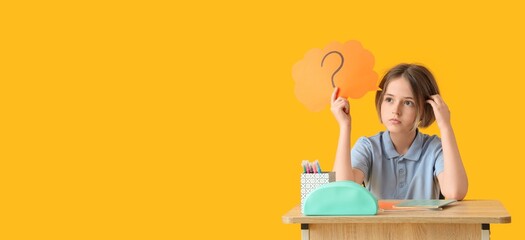 Thoughtful schoolboy holding speech bubble with question mark while sitting at desk on yellow background with space for text