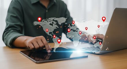 Close up of a person's hands interacting with a digital world map projected onto a desk with location pins and glowing elements on a laptop and tablet