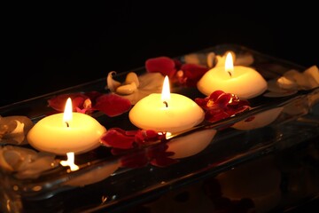 Burning candles and orchid flowers in glass container of water on mirror table against black background, closeup