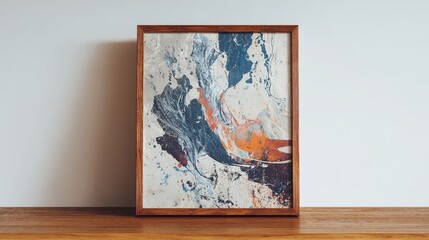 A framed abstract painting featuring swirling, marbled textures in shades of white, blue, orange, and grey, rests on a light wood surface against a white wall