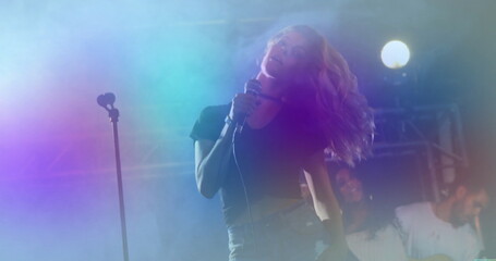 Performing female singer holding handheld microphone on concert stage, with fog and electric guitar
