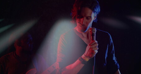 Singing singer holding wired microphone on stage, guitarist strumming guitar under spotlights