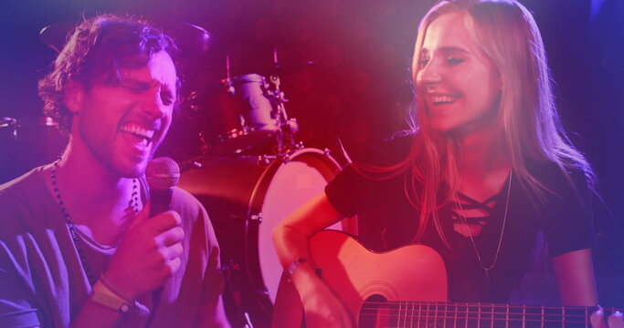Singing singer holding microphone as guitarist strumming guitar on stage, with drum kit and lights