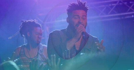 Singing singer wearing dark shirt holding mic gesturing on stage with bass guitar truss and lights