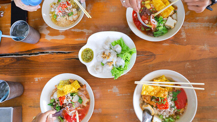 Friends gather around a rustic wooden table to share bowls of colorful noodle dishes, accompanied...
