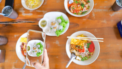 A person holds up a small plate of colorful Thai dishes while a spread of various bowls with noodles, soup, and fresh vegetables sits on the table. Friends enjoy lunch together