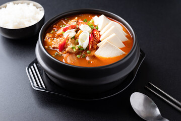 Korean food (Kimchi Jjigae), Kimchi soup with tofu, pork, scallion and chili in Korean stone bowl eating with cooked rice on black background