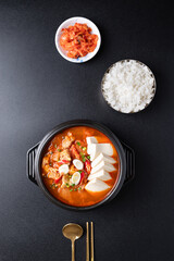 Top view of Korean food (Kimchi Jjigae), Kimchi soup with tofu, pork, scallion and chili in Korean stone bowl eating with cooked rice and kimchi on black background