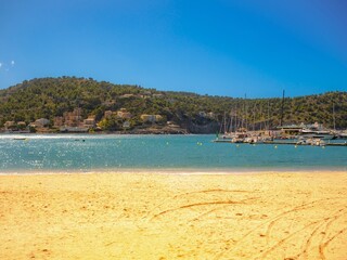 Golden sand at Platja des Traves, Port de Soller—turquoise bay, moored yachts and the Serra de Tramuntana backdrop in Mallorca, Spain; classic Balearic summer under a clear blue sky.