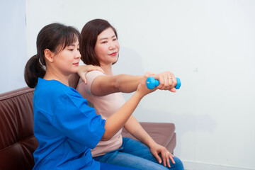 Young Asian nurse assisting middle-aged female patient on a couch doing physical therapy, focusing...