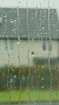 Rainy day ambiance through window, droplets sliding down, soft focus house exterior behind glass