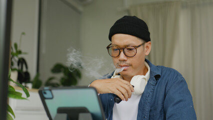 Asian man sitting at his desk and working on a laptop while smoking an electronic cigarette.