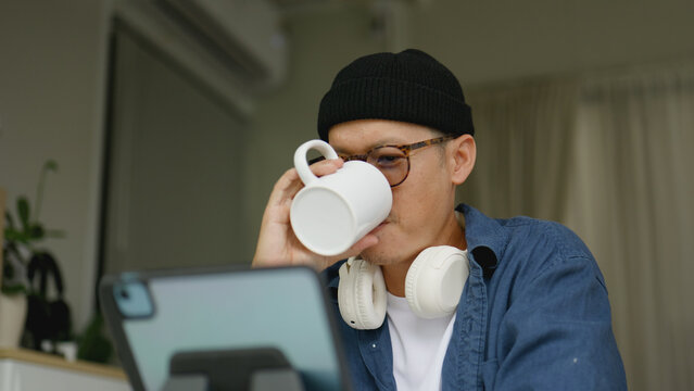 Asian man working from home on his tablet while sipping coffee.