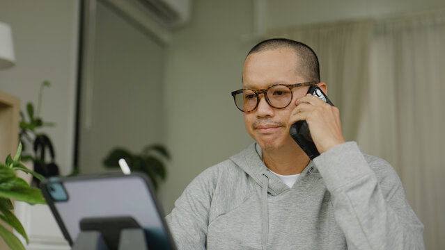 Asian man talking on his smartphone while working on a tablet.