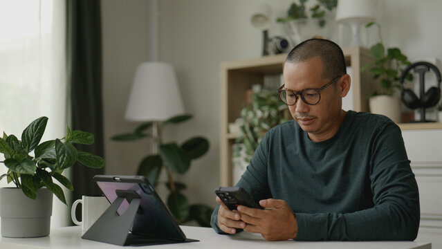 Asian man multitasking with his smartphone while checking information on a tablet.