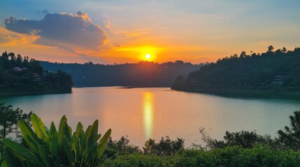 Naklejka premium 23.Scenic lakeside sunset with the vibrant golden sun dipping below the horizon, its light reflecting on the glassy lake water, framed by dense greenery and gentle hills, all under a fading blue sky.