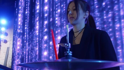Female drummer musician passionately performing on stage, striking cymbals with vibrant red drumsticks against dramatic purple LED lighting backdrop, capturing intense musical energy - Powered by Adobe