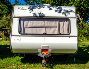 Rear view of a white vintage caravan parked on green grass under sunny skies, surrounded by lush trees, ready for a serene summer holiday adventure.