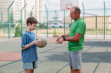 Father teaches his son how to play basketball. The boy throws the ball into the hoop at the...