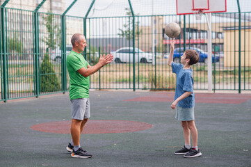 Father teaches his son how to play basketball. The boy throws the ball into the hoop at the playground. Generation. Sports. Hobbies