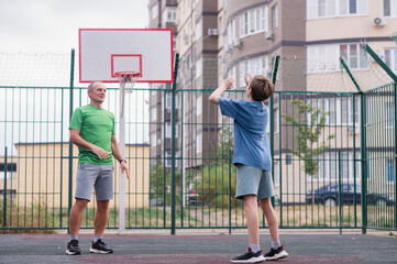 Father teaches his son how to play basketball. The boy throws the ball into the hoop at the playground. Generation. Sports. Hobbies