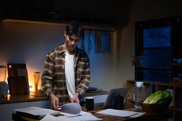 Man working late at home, reviewing paperwork on kitchen table with laptop