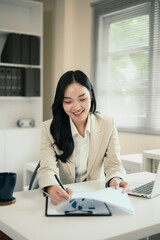 Smiling businesswoman writing notes while reviewing financial reports at office desk
