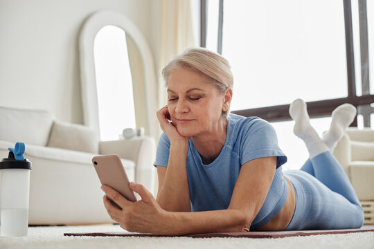 Mature Caucasian woman lying on floor using smartphone, resting chin on hand, relaxing in bright room, exercise mat and water bottle nearby, engaging with mobile device - Powered by Adobe