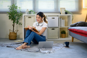 Young woman sitting on floor at home, writing in notebook with laptop and coffee nearby