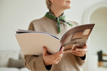 Caucasian senior woman holding open magazine with interior design images, standing indoors with...