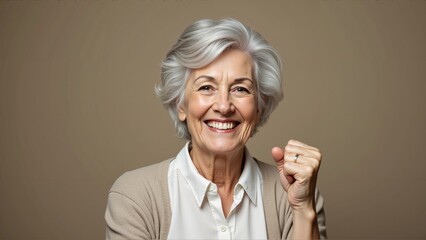 Smiling senior woman with gray hair raising fist in confidence, wearing white shirt and beige cardigan, joyful and positive expression against neutral background, indoor studio portrait with natural l