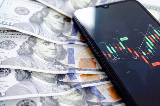 Smartphone con gr&aacute;ficos finacieros de velas japonesas sobre billetes de 100 d&oacute;lares. Inversiones y trading.   