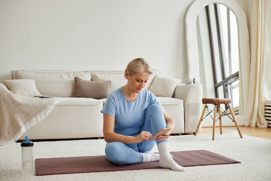 Caucasian senior woman sitting on yoga mat using smartphone during home workout session, exercising in living room with water bottle nearby, mirror and sofa in background