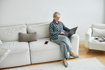 Senior Caucasian woman sitting on sofa using laptop, wearing glasses and smiling while working...