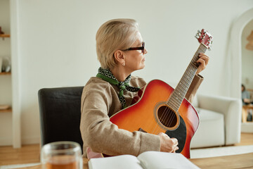 Senior Caucasian woman wearing sunglasses playing acoustic guitar while sitting indoors, holding...