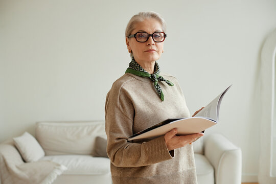 Portrait of senior Caucasian woman wearing glasses standing indoors holding open book looking at camera with confident expression, short gray hair visible, neutral background