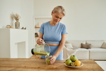 Adult Caucasian woman pouring green smoothie from blender into glass, standing at wooden table with...