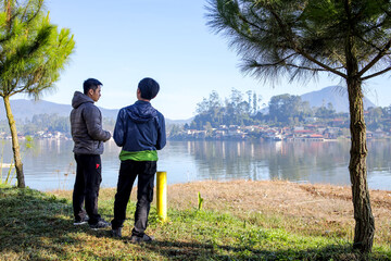 Two Men Standing By Lake Admiring The Scenic View Of The Water Distant Village And A Mountain
