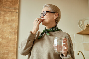 Senior Caucasian woman wearing glasses holding glass of water taking medication standing in kitchen...