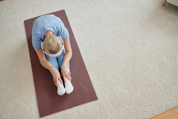 Senior Caucasian woman sitting on yoga mat stretching forward with hands touching feet practicing...