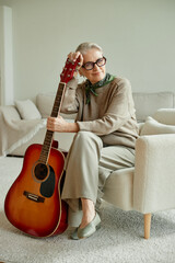 Senior Caucasian woman sitting on chair holding acoustic guitar, wearing glasses and scarf, smiling...