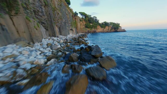 FPV drone flies low above the sea at blue hour, skimming water and cliffs, passing stones, a road, a person, turning around them and continuing along the coastline.