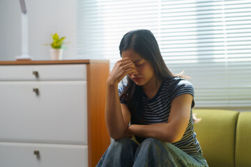 Sad young woman sitting on sofa at home, covering her face with her hand and suffering from a bad...